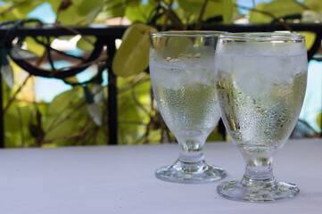 Two Misty Glasses of Cold Clean Water with Ice are on White Table - on Green Leaves and Blue Air Background, Copy Space. Fresh Drink of Hot Summer.