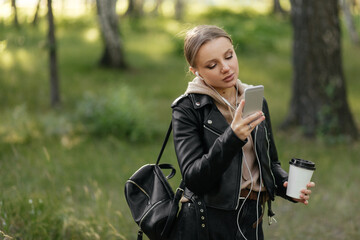 A beautiful woman in a leather jacket and headphones walks in the park, checks mail on the phone and drinks coffee. Walks in the park.