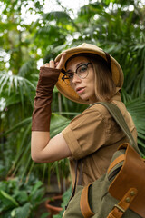 Woman botanist dressed in safari style in greenhouse. Naturalist in khaki clothes, leaver gloves with backpack walks in the rainforest surrounded by palms, looking at camera. Jungle tourist