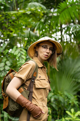 Woman botanist dressed in safari style in greenhouse. Naturalist in khaki clothes, leaver gloves with backpack walks in the rainforest surrounded by palms, looking at camera. Jungle tourist