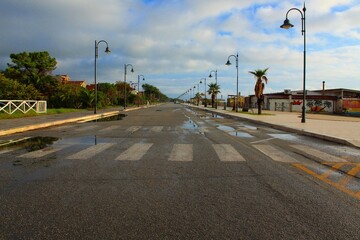 Fototapeta na wymiar waterfront street is wet after early morning rain, Calabria ( Ardore) , puddles along the empty road and crosswalk, perspective with rows of street lamps along the sidewalks, cloudy blue sky