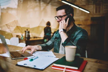 Prosperous male freelancer talking on telephone while doing remote job in cafe interior, cheerful hipster guy watching funny video on laptop computer joking with friend on cellphone conversation