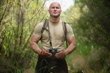 Adventurer man with a binoculars in the hands is standing in the grass on green forest background.