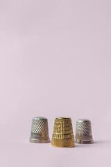 Vertical image of a triangular composition of three thimbles, two types, and one gold on a pink background. Sewing items.