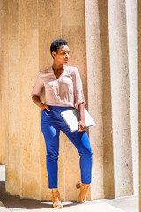 Young African American Businesswoman with short afro hair, wearing light color V neck shirt, blue pants, brown sandal high heels, holding laptop computer, standing by column in New York City, frowned.