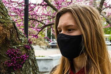 Portrait of young woman in protective medical face mask with flowers near blooming tree in spring time. Coronavirus COVID-19 epidemic protection. Allergic to flowering in spring concept.