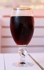 A glass of cold dark beer on a white wooden table in cafe. The glass is covered with small water droplets. Concept of hot summer thirst quenching, relaxation and fun, drinking beverages