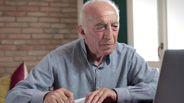 A senior man sitting at a table and using a laptop, taking some notes, at home. Slow camera movement.