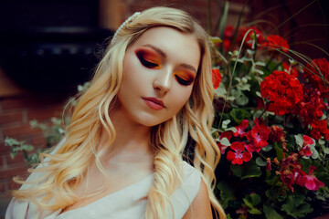 Portrait of a pensive blonde near red flowers. Professional makeup, red smoky eyes. Girl with a cute pretty face.