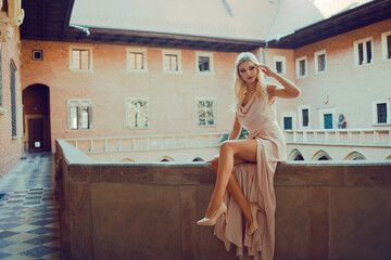 A beautiful girl with long legs is sitting on the balcony in the castle. Princess on the balcony on vacation. The woman in the historic city. Model in a chic long pink dress with heels