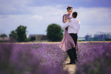 Young couple in love hugging and walking in a lavender field on summer cloudy day. girl in a luxurious purple dress and with hairstyle