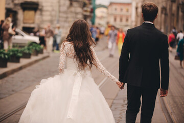 Happy newlyweds couple on a walk in old European town street, gorgeous bride in white wedding dress together with handsome groom. rear view