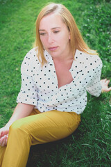 Obraz premium beautiful middle age woman sitting on a grass. thoughtful girl wearing trendy polka dot blouse and yellow pants. calm and pensive woman. 