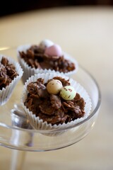 Corn flakes coated chocolate cake with chocolate mini Easter eggs on top in a paper cake case displayed in a glass cake stand,