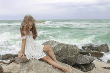 Fototapeta na wymiar Little beautiful girl child at the sea under the blue sky sitting on the stones during a storm