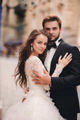 Naklejka premium Happy newlywed couple hugging and kissing in old European town street, gorgeous bride in white wedding dress together with handsome groom. wedding day