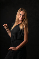 Playful beautiful girl with long blonde hair in a dark dress on a black background portrait in a half-turn, holding a belt and smiling.