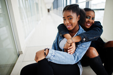 Two african woman friends in jeans jacket sitting indoor together.