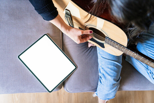 Woman playing guitar sitting on her couch at home and learning with online lessons with a digital tablet with a digital tablet with a blank screen from above