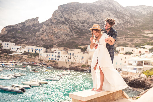 Cheerful traveling couple standing on rocky hill and embracing on background of majestic seascape with boats and residential houses