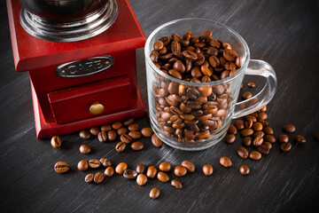 Retro coffee grinder and glass transparent cup full of brown coffee beans and grains around on dark...
