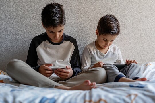 Brothers sitting on cozy bed and watching cartoons on cellphones while relaxing at home
