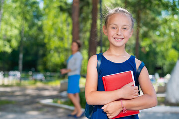 Portrait of a diligent schoolgirl with a backpack and a book goes to first grade. Mom sends a happy daughter to elementary school.