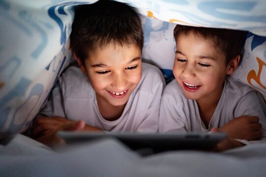 Positive siblings in pajamas hiding under blanket and enjoying interesting cartoon during daytime at home