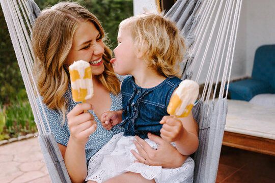 Cheerful mother and daughter hugging in hammock on terrace with tasty ice lollies and enjoying summer together