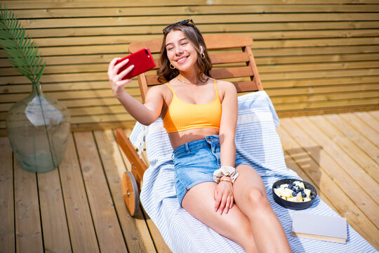 From above positive female in shorts and bra lying on deck chair on sunny day and taking photo on camera of cellphone