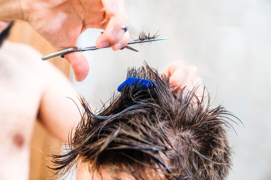 Unrecognizable male making haircut to guy using scissors against blurred interior of light bathroom at home