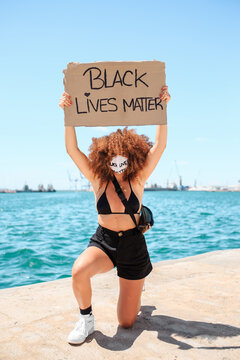 Ethnic female activist in medical mask and with black lives matter placard taking knee in solidarity during peaceful protest in city and looking at camera