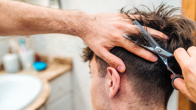 Back view of unrecognizable male making haircut to guy using scissors against blurred interior of light bathroom at home