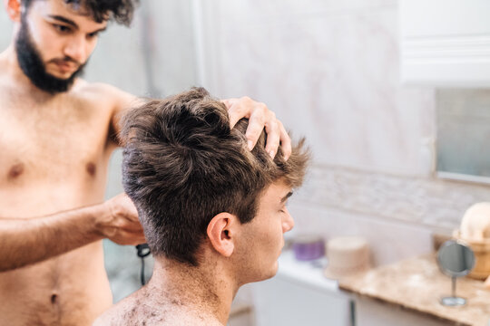 Male with hair trimmer cutting hair of guy in contemporary bathroom at home