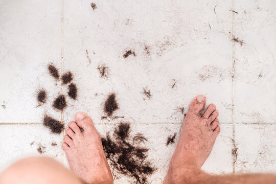 Barefoot unrecognizable male sitting on chair among pile of trimmed dark hair in light modern bathroom at home