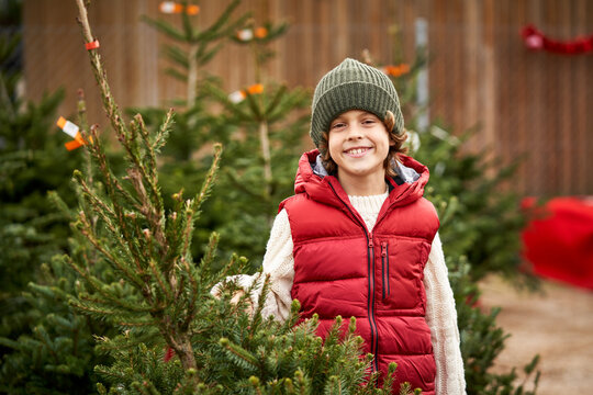 Beautiful blond boy with green wool hat, red vest, white pullover, blue pants and yellow boots choosing his Christmas tree