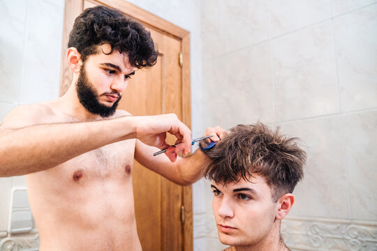 Male making haircut to guy using scissors against blurred interior of light bathroom at home