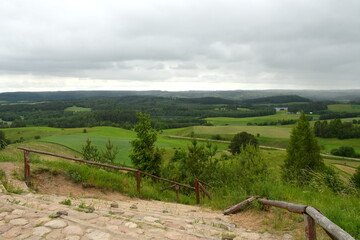 A view from the top of a tall mountain with stone stairs connected to wooden handles visible and a dense forest or moor, meadows, fields, and shrubbery visible below the hilltop