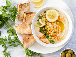 Hummus with whole grains of hummus on a white plate with lemon, parsley and pita on a white-grey background.
