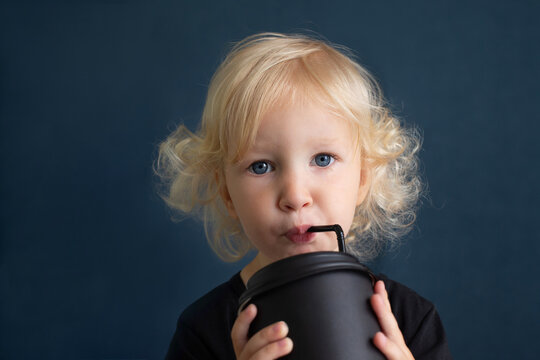 Oddler Girl With Blond Curly Hair And Blue Eyes Drinking From Big Black Reusable Bamboo Cup Using Straw. Girl Is Looking At Camera. Portrait Is Made On Dark-blue Background.