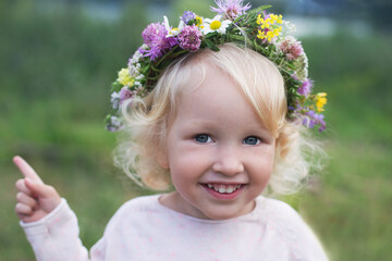Portraite of small pretty smiling girl with blond curly hair and blue eyes wearing wild flowers wreath. Summer mood and carefree childhood concept.