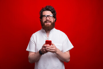 Young man wearing glasses is looking at the camera while holding phone over red background