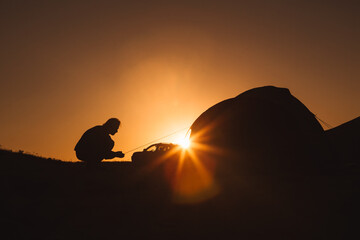 A man is silhouetted against the orange sky of sunset or dawn. A man sets up a camp in the mountains near the car. Journey.
