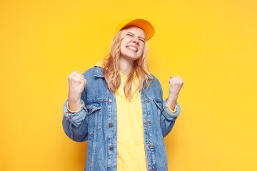 crazy girl in a denim jacket and a cap rejoice at success on a yellow background, winner girl enjoy victory