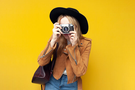 girl in autumn clothes with a backpack holds a camera and takes a photo on a yellow isolated background