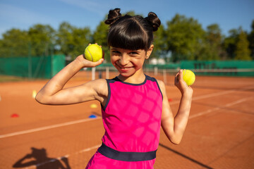 a smiling girl posing on the tennis court with two tennis balls