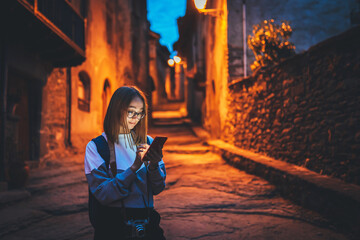 tourist girl using mobile phone with glasses reflection of screen light of street old historical city at night, woman plans walk architectural sights of old town looks at online map in cellphone