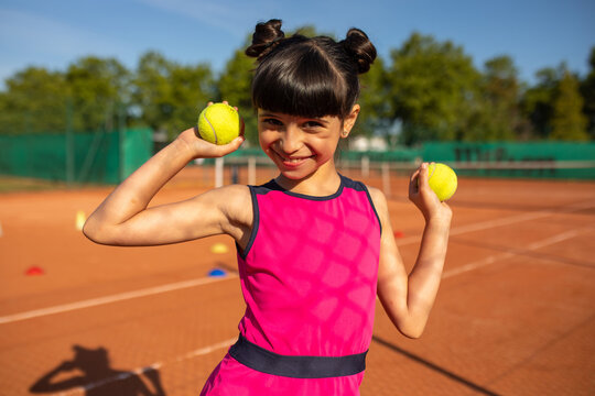 a smiling girl posing on the tennis court with two tennis balls