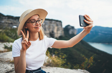 smiling female traveler in summer hat communication  by video internet call on smartphone walking on top of mountain landscape, fun female tourist takes photo selfie on mobile phone on nature outdoors