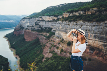 young blonde girl takes picture on retro photo camera on background panorama horizin mountain and river landscape, hipster tourist enjoys hobby of photographer relax in summer nature